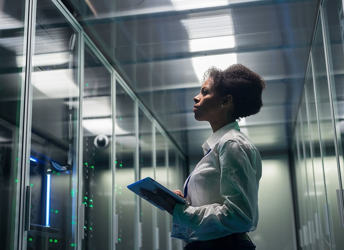 Business Insurance - Portrait of a Young Female Engineer Holding a Tablet Looking at a Room of Servers Through the Glass