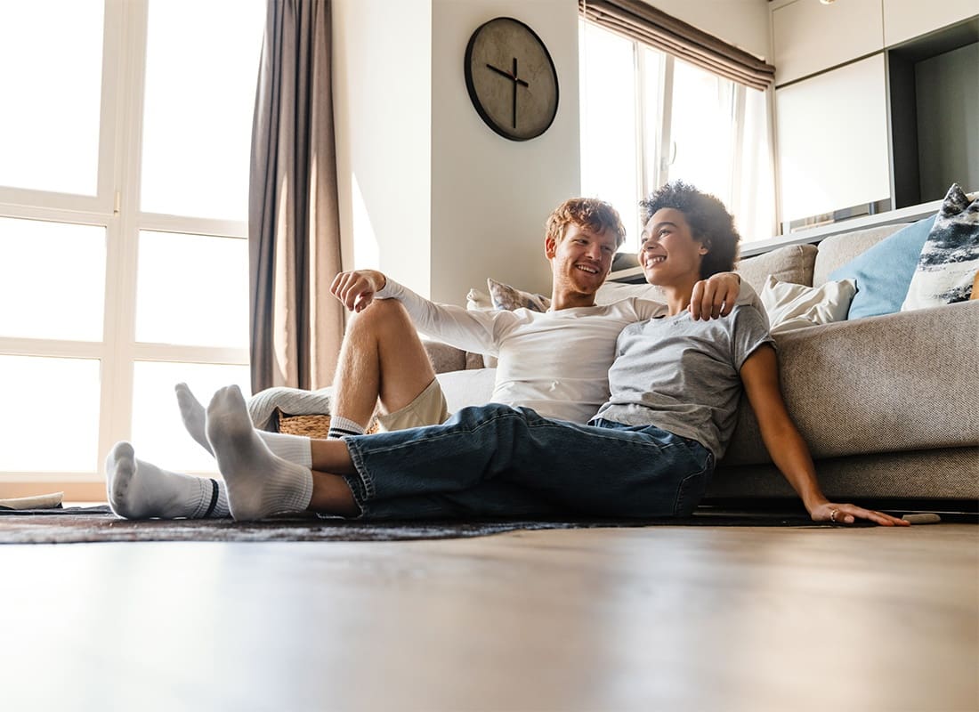 Personal Insurance - Portrait of a Cheerful Young Diverse Married Couple Sitting on the Floor Next to the Sofa in the Living Room